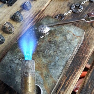 May include: A close-up of a silver cufflink with a detailed design being heated with a blowtorch on a metal surface. The cufflink is being held in place with tweezers.