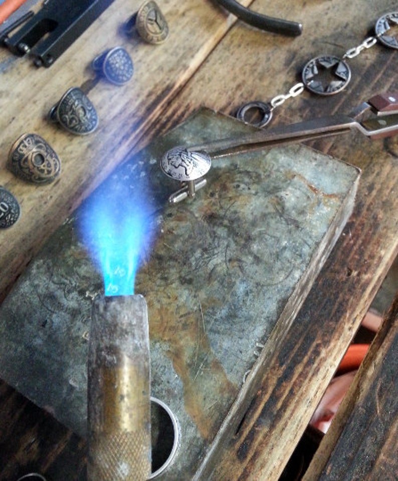 May include: A close-up of a silver cufflink with a detailed design being heated with a blowtorch on a metal surface. The cufflink is being held in place with tweezers.