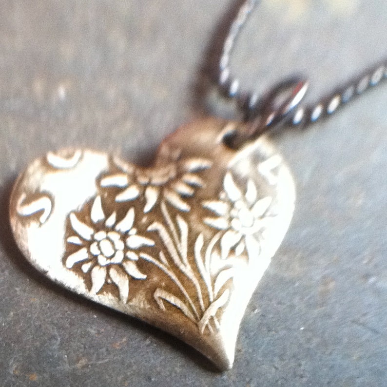 K&ouml;nnte beinhalten: Eine silberne herzf&ouml;rmige Halskette mit einem filigranen Design aus Edelwei&szlig;-Blumen. Der Anh&auml;nger ist an einer schwarzen Kette befestigt.