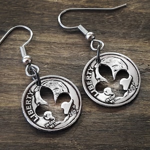 May include: Silver-colored coin earrings with a fleur-de-lis design cut out of the center. The earrings are made from quarters dated 2002, with silver-colored fish hook style ear wires. The earrings are displayed on a wooden surface.