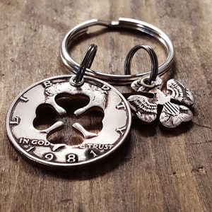 May include: A silver-colored keychain featuring a cut-out shamrock design on a coin dated 1981, with the words "IN GOD WE TRUST" visible. An additional charm depicts an eagle. The keychain is on a wooden surface.