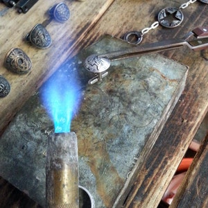 Op de afbeelding: Een close-up van een zilveren manchetknoop met een gedetailleerd ontwerp dat wordt verwarmd met een brander op een metalen oppervlak. De manchetknoop wordt met een pincet op zijn plaats gehouden.
