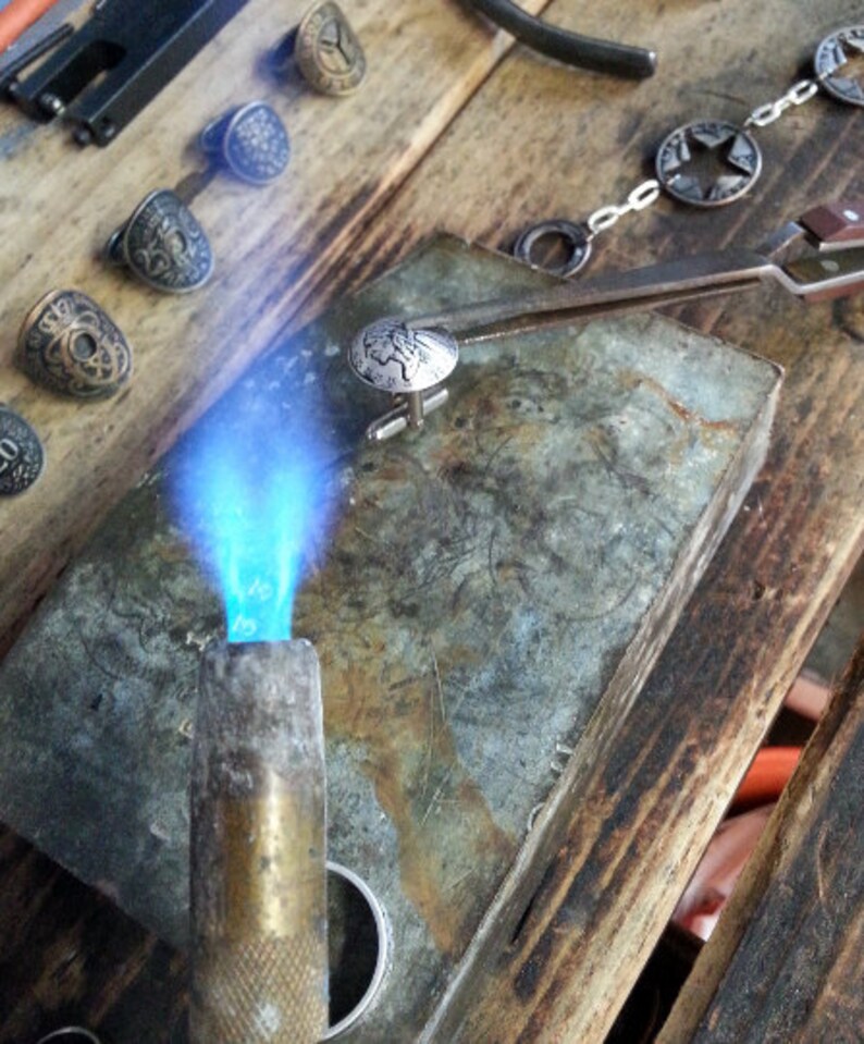 Op de afbeelding: Een close-up van een zilveren manchetknoop met een gedetailleerd ontwerp dat wordt verwarmd met een brander op een metalen oppervlak. De manchetknoop wordt met een pincet op zijn plaats gehouden.