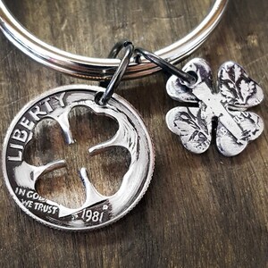 May include: A silver-colored keychain featuring a four-leaf clover charm and a coin with a clover cutout. The coin is dated 1981 and has the words "LIBERTY" and "IN GOD WE TRUST" engraved on it. The keychain is attached to a silver ring.