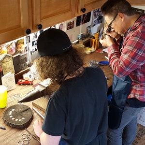 May include: Two people working on a jewelry making project. One person is using a magnifying glass to examine a piece of jewelry, while the other person is using a hammer to shape metal. There are tools and supplies on the table, including a small red toy truck.