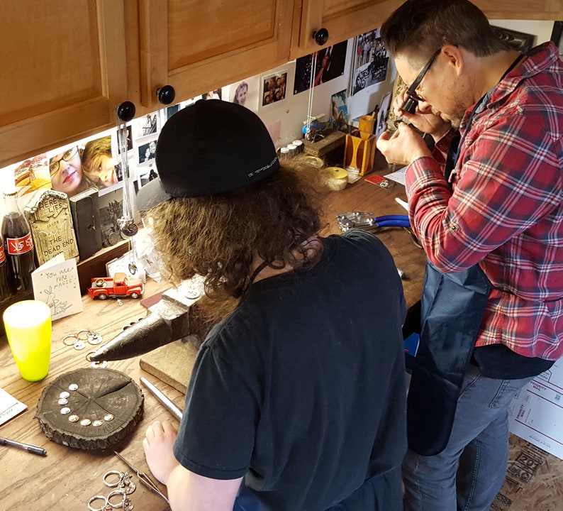 May include: Two people working on a jewelry making project. One person is using a magnifying glass to examine a piece of jewelry, while the other person is using a hammer to shape metal. There are tools and supplies on the table, including a small red toy truck.