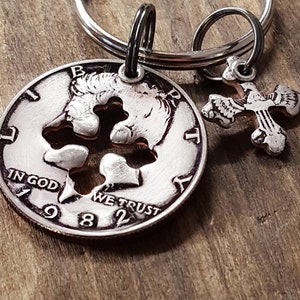 May include: A silver-colored keychain featuring a cut-out coin with a cross design and a small cross charm. The coin is dated 1982 and has the words "IN GOD WE TRUST". The keychain is attached to a silver ring.