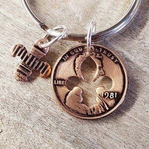 May include: A silver keychain with a copper-colored cross charm and a cut-out penny from 1981. The penny has a cut-out design and the words "IN GOD WE TRUST" and "LIBERTY" are visible.
