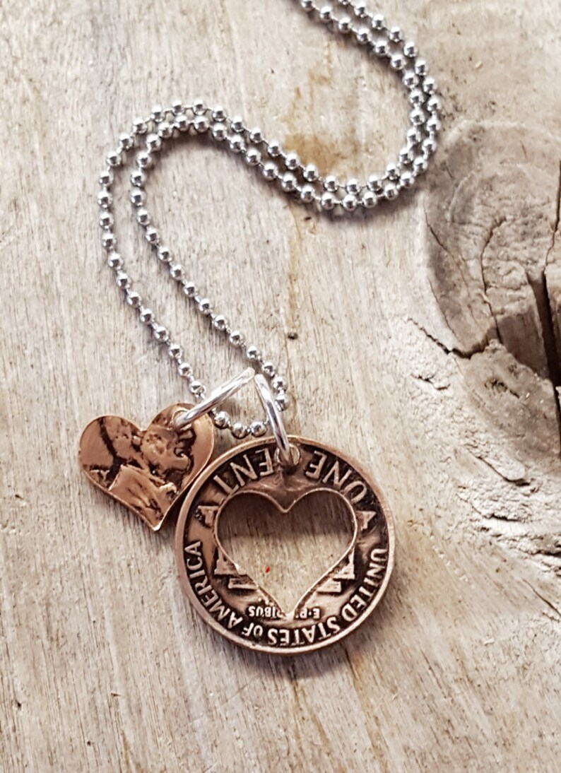 Op de afbeelding: Een zilveren ketting met een koperen hanger met een hart dat uit een cent-munt is geknipt. De munt heeft de tekst "ONE CENT" en "UNITED STATES OF AMERICA" met het jaar "1985" en "E PLURIBUS UNUM". Een kleinere koperen hartbedel is aan de munt bevestigd.