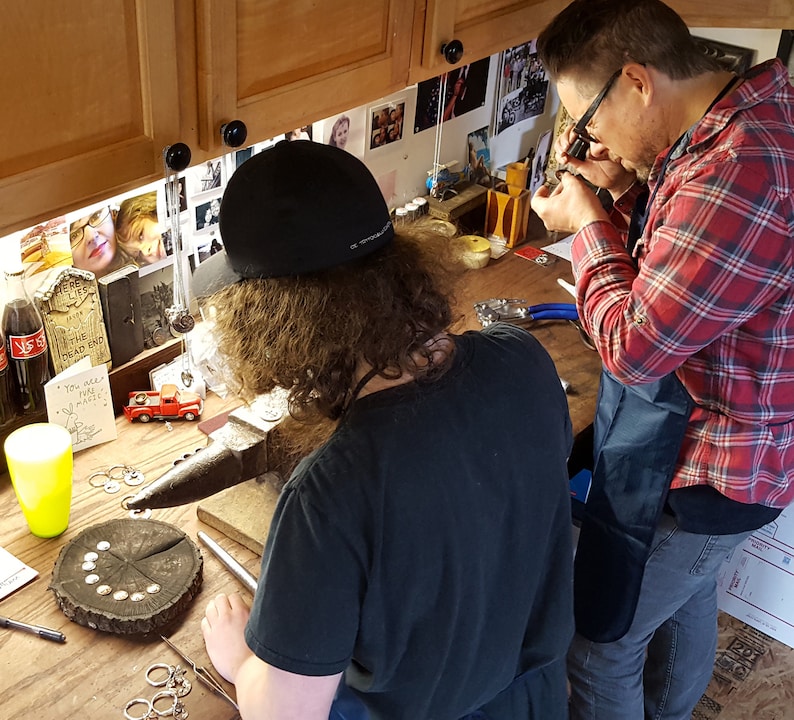 Op de afbeelding: Twee mensen werken aan een juweelmaakproject. Een persoon gebruikt een vergrootglas om een ​​juweel te onderzoeken, terwijl de andere persoon een hamer gebruikt om metaal te vormen. Er zijn gereedschappen en benodigdheden op de tafel, waaronder een kleine rode speelgoedtruck.