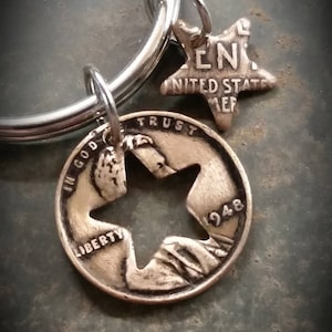May include: A silver-toned keychain featuring a cut-out coin with a star-shaped hole and a star charm. The coin is dated 1948 and has the word "LIBERTY" visible. The star charm has the text "UNITED STATES OF AMERICA".