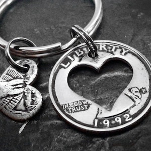 May include: A silver-toned keychain featuring a heart-shaped cutout coin from 1992, with the words "LIBERTY" and "IN GOD WE TRUST" visible. A small heart-shaped charm with an engraved design hangs next to the coin. The keychain is attached to a silver ring.