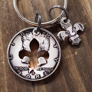 May include: A silver-colored keychain featuring a cut-out fleur-de-lis design on a coin dated 1988, with the words "IN GOD WE TRUST". A smaller fleur-de-lis charm hangs next to it. The keychain is on a wooden surface.