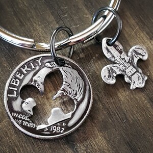 May include: A silver-colored keychain featuring a cut-out coin with the word "LIBERTY" and the year 1982, along with a fleur-de-lis charm. The keychain is attached to a silver ring. The background is a wooden surface.