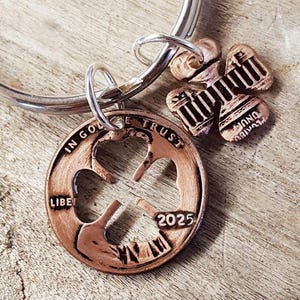 May include: A silver key chain with a copper penny charm featuring a four-leaf clover cutout. The penny has the inscription "IN GOD WE TRUST" and "LIBERTY" and the year "2025". A smaller copper charm shaped like a four-leaf clover with the inscription "E PLURIBUS UNUM" is attached to the key chain.