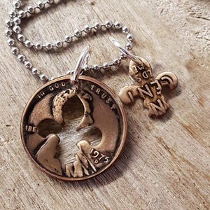 May include: A copper-colored pendant necklace featuring a cut-out design on a coin from 1975, with the words "In God We Trust" visible. A smaller, cream-colored charm with the word "GENUINE" is also included. The necklace has a silver ball chain.