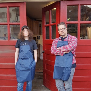 May include: Two people wearing blue aprons stand in front of a red garage door. The person on the left is wearing a black t-shirt and jeans. The person on the right is wearing a red and black plaid shirt and jeans.
