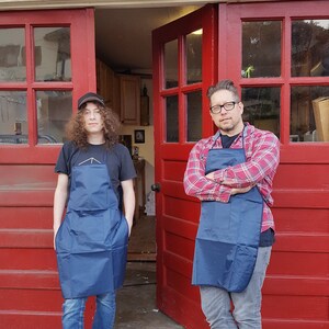May include: Two people wearing blue aprons stand in front of a red garage door. The person on the left is wearing a black t-shirt and jeans. The person on the right is wearing a red and black plaid shirt and jeans.