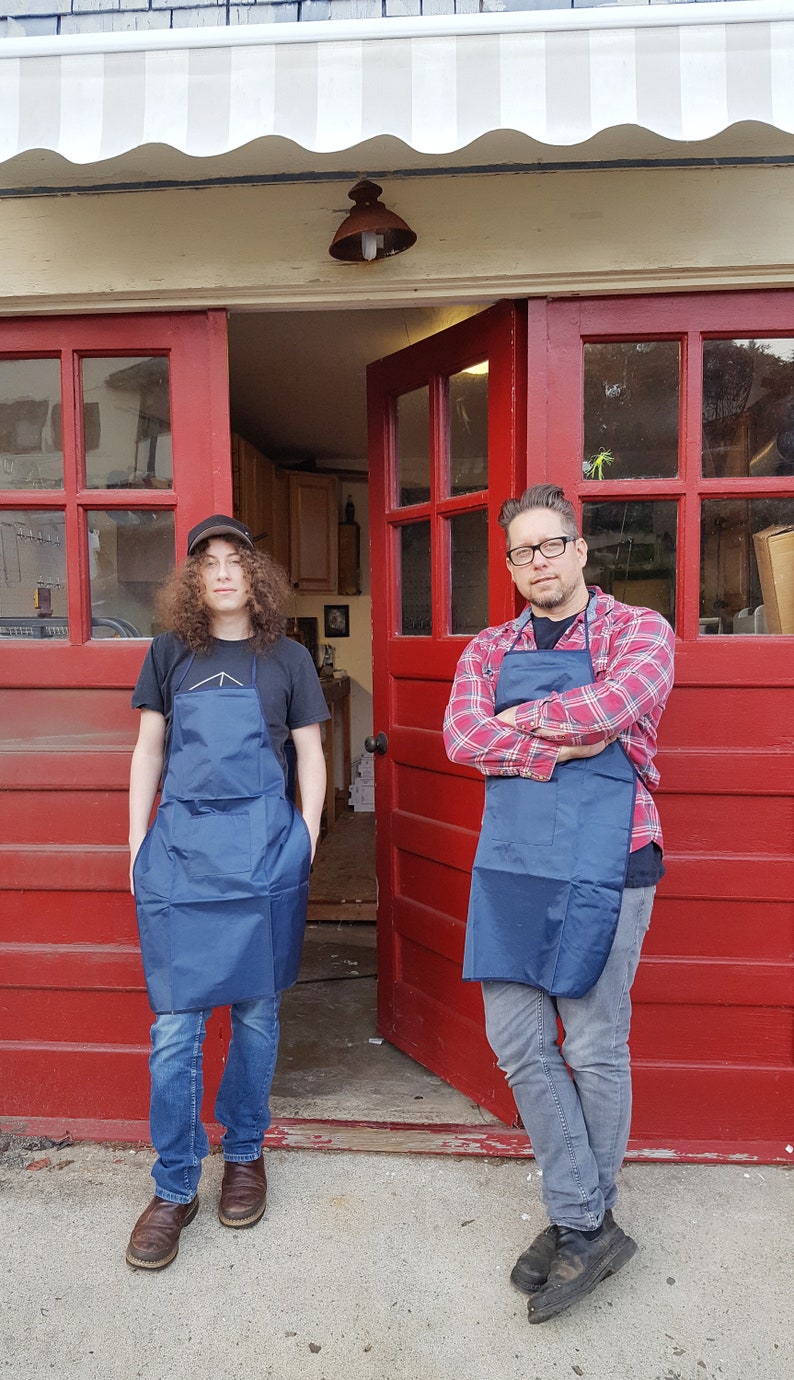May include: Two people wearing blue aprons stand in front of a red garage door. The person on the left is wearing a black t-shirt and jeans. The person on the right is wearing a red and black plaid shirt and jeans.