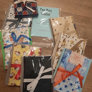 May include: A collection of fabric tea bag wallets in various patterns, including musical notes, chickens, and patchwork designs. Each wallet is packaged in clear plastic and tied with a ribbon. A blue card reads "Tea Bag Wallet".