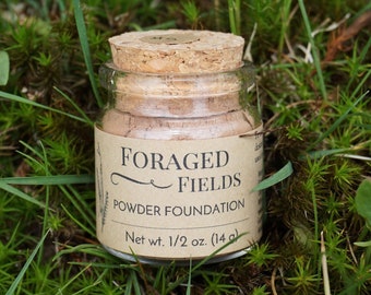 Op de afbeelding: Een klein glazen potje poederfoundation met een kurken deksel, met het label "FORAGED FIELDS". Het label geeft ook "Powder Foundation" en "Netto gewicht 1/2 oz. (14 g)" aan. De pot staat op een bed van groen gras en mos.