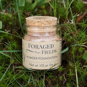 May include: A small glass jar of powder foundation with a cork lid, labeled "FORAGED FIELDS." The label also indicates "Powder Foundation" and "Net wt. 1/2 oz. (14 g)." The jar sits on a bed of green grass and moss.