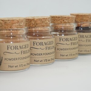 May include: A collection of small glass jars with cork stoppers, filled with powder foundation. Each jar has a label that reads "FORAGED FIELDS POWDER FOUNDATION" and "Net wt. 1/2 oz. (14g)". The jars are arranged in a row against a white background.