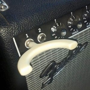 May include: Close-up of a black and silver guitar amplifier. The amp features control knobs labeled Treble, Bass, and Volume. A white handle is visible, along with the brand name Fender in a silver script.