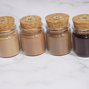 May include: Five small glass jars with cork stoppers, each filled with a different shade of powder. The powders range in color from light beige to dark brown. Each cork has a small sticker with a number on it.