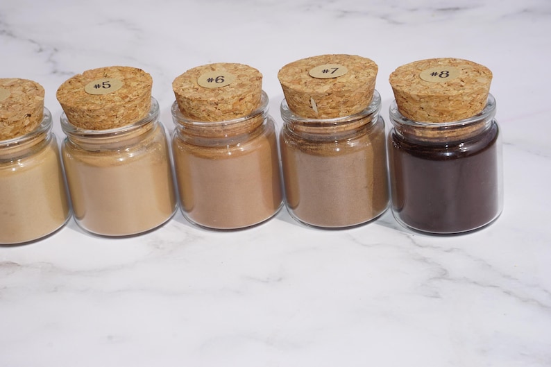 May include: Five small glass jars with cork stoppers, each filled with a different shade of powder. The powders range in color from light beige to dark brown. Each cork has a small sticker with a number on it.