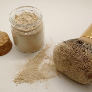 May include: A makeup brush with a wooden handle and gold ferrule next to a jar of beige powder and a cork. The brush has a large, soft head. A small amount of powder is spilled on the surface.