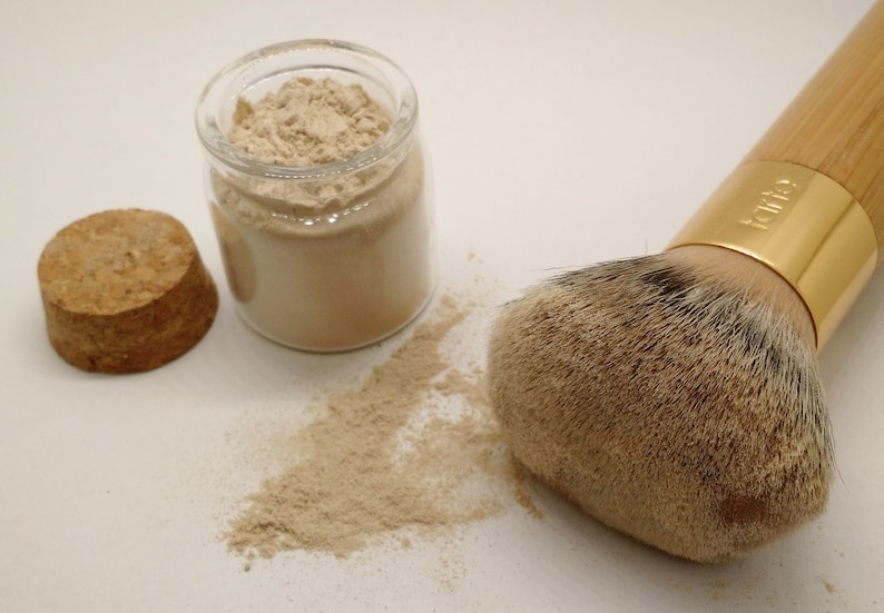 May include: A makeup brush with a wooden handle and gold ferrule next to a jar of beige powder and a cork. The brush has a large, soft head. A small amount of powder is spilled on the surface.
