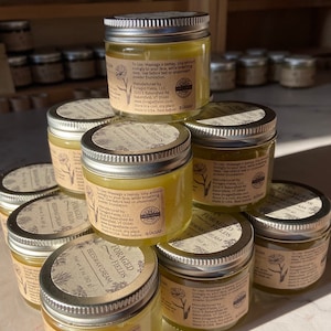 May include: A stack of small glass jars filled with a yellow cream. The jars have silver lids and are labeled "Foraged Fields Beeswax Cream". The jars are stacked on a wooden surface.