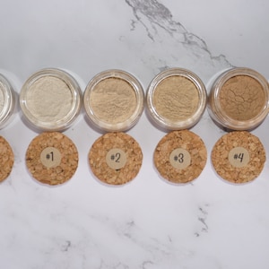 May include: A collection of six small, clear jars filled with varying shades of beige powder, each with a cork lid. The lids are labeled with numbers from 0 to 5. The jars are arranged in a row on a white marble surface.