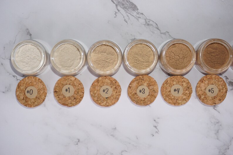 May include: A collection of six small, clear jars filled with varying shades of beige powder, each with a cork lid. The lids are labeled with numbers from 0 to 5. The jars are arranged in a row on a white marble surface.