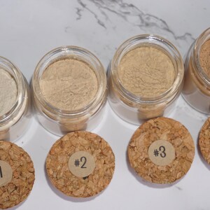 May include: Four small glass jars filled with different shades of powder, ranging from light beige to brown. Each jar has a cork lid with a numbered sticker. The jars are arranged in a row.