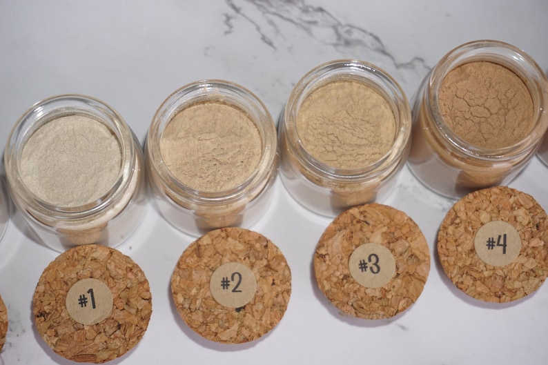 May include: Four small glass jars filled with different shades of powder, ranging from light beige to brown. Each jar has a cork lid with a numbered sticker. The jars are arranged in a row.