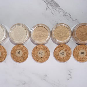 May include: A selection of six small, clear jars filled with various shades of powder. Each jar is paired with a cork lid labeled with a number from 0 to 5. The powders range in color from light beige to medium brown.