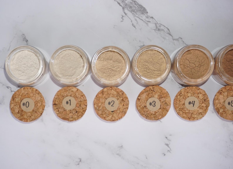 May include: A selection of six small, clear jars filled with various shades of powder. Each jar is paired with a cork lid labeled with a number from 0 to 5. The powders range in color from light beige to medium brown.
