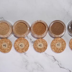 May include: A collection of six small, clear jars filled with various shades of brown powder, each with a cork lid. The lids are numbered from #3 to #8. The powders range in color from light beige to dark brown.