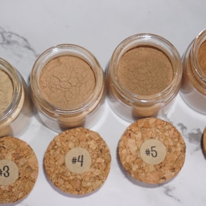 May include: A collection of small glass jars filled with various shades of brown powder, each with a cork lid labeled with a number. The powders range in color from light tan to dark brown. The jars are arranged on a white marble surface.