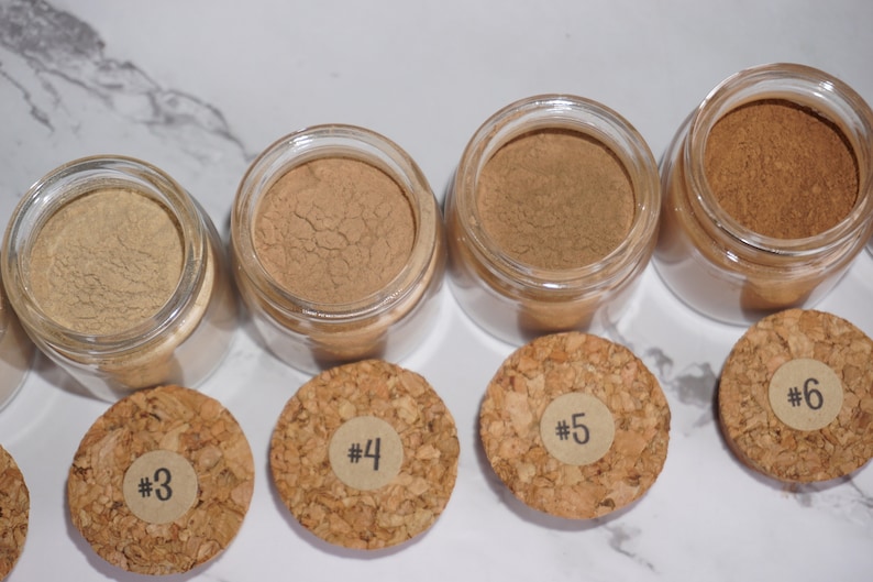 May include: A collection of small glass jars filled with various shades of brown powder, each with a cork lid labeled with a number. The powders range in color from light tan to dark brown. The jars are arranged on a white marble surface.