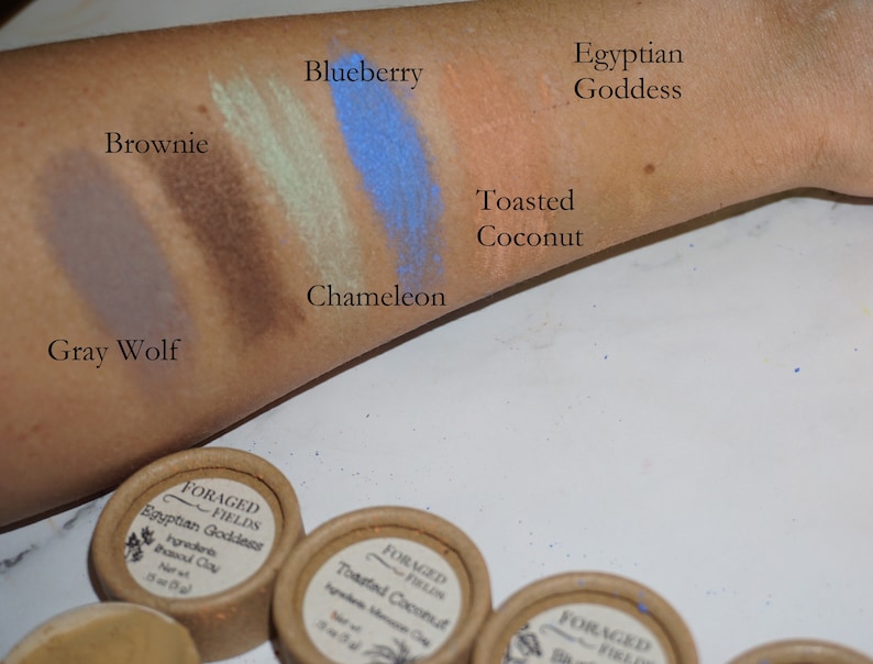 May include: Swatches of eyeshadow on a person's arm. The colors are: Gray Wolf, Brownie, Chameleon, Blueberry, Toasted Coconut, and Egyptian Goddess. The eyeshadow is in small containers labeled "Foraged Fields".