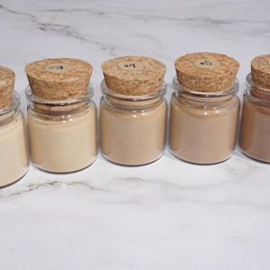 May include: A row of small glass jars filled with various shades of beige and brown powder. Each jar has a cork stopper with a small, round label. The jars are arranged on a white marble surface.
