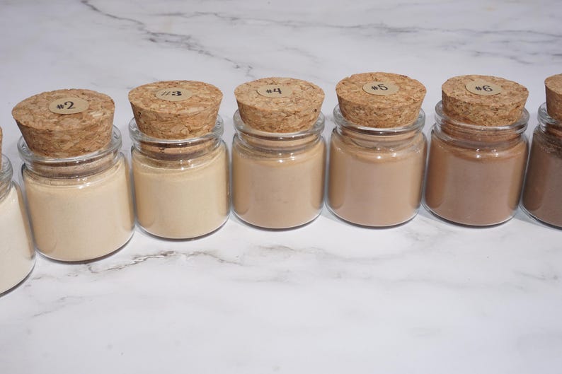 May include: A row of small glass jars filled with various shades of beige and brown powder. Each jar has a cork stopper with a small, round label. The jars are arranged on a white marble surface.