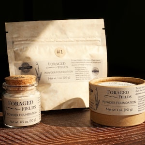 May include: Three containers of Foraged Fields Powder Foundation. A small glass jar with a cork lid, a round cardboard container, and a paper pouch. The containers are labeled with the product name and net weight: 1/2 oz (10 g) and 1 oz (20 g).