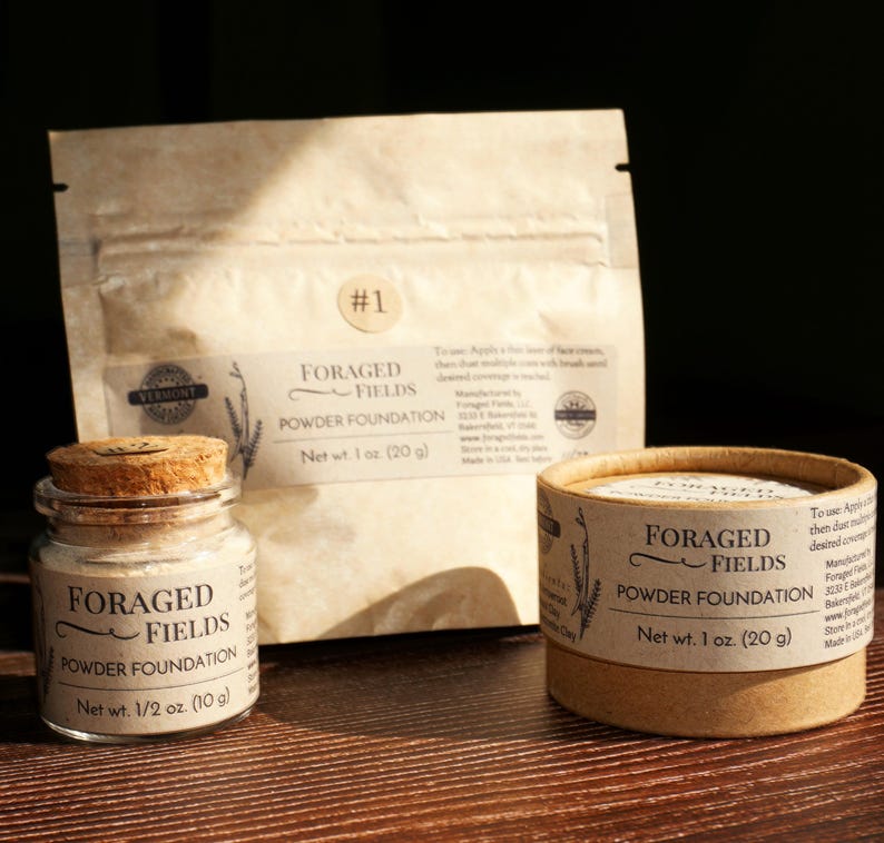 May include: Three containers of Foraged Fields Powder Foundation. A small glass jar with a cork lid, a round cardboard container, and a paper pouch. The containers are labeled with the product name and net weight: 1/2 oz (10 g) and 1 oz (20 g).