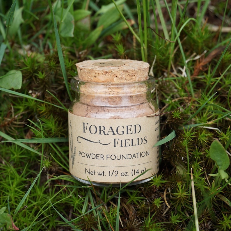 May include: A small glass jar of powder foundation with a cork lid, labeled "FORAGED FIELDS." The label also indicates "Powder Foundation" and "Net wt. 1/2 oz. (14 g)." The jar sits on a bed of green grass and moss.