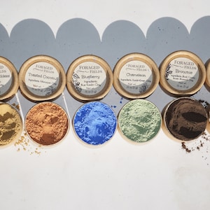 May include: Six small round containers of different colored clay powders, labeled with the names "Egyptian Goddess", "Toasted Coconut", "Blueberry", "Chameleon", "Brownie", and "Limited Edition".