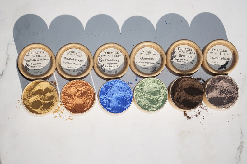 May include: Six small round containers of different colored clay powders, labeled with the names "Egyptian Goddess", "Toasted Coconut", "Blueberry", "Chameleon", "Brownie", and "Limited Edition".
